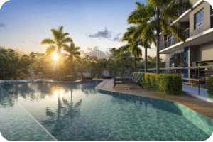 Área de lazer de um apartamento de luxo em Fortaleza. A imagem, registrada ao pôr do sol, mostra uma piscina de borda infinita com revestimento em tom azul-turquesa, um deck de madeira com espreguiçadeiras, palmeiras e muito verde ao fundo, transmitindo uma atmosfera de relaxamento.