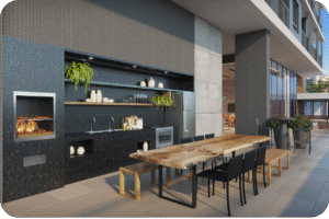 Área gourmet externa e coberta de um apartamento de luxo em Fortaleza. O espaço possui uma churrasqueira moderna, bancadas escuras, eletrodomésticos em aço inox e uma grande mesa de madeira rústica e sólida, com cadeiras pretas, ideal para convivência e lazer.